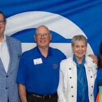 four people, all wearing blue, pose for photo in front of GVSU banner at celebration event